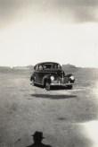 Black and white photograph of an early car in an empty landscape with the photographer's silhouette on the ground