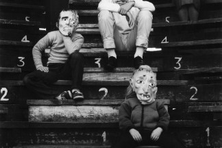 Black-and-white photograph of four figures in Halloween masks seated on a numbered riser.