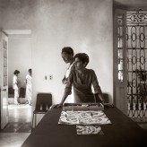 Sepia toned photograph of a woman leaning on a table in front of a boardgame and another woman staring directly into the camera behind her.