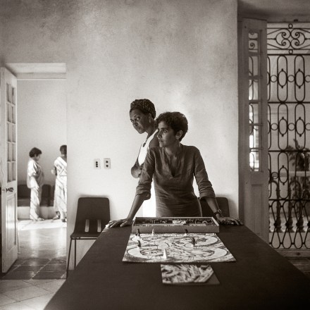 Sepia toned photograph of a woman leaning on a table in front of a boardgame and another woman staring directly into the camera behind her.