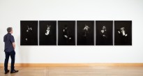 Color image of a figure viewing six black and white photographs depicting six different animals in suits on white gallery wall