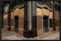 A color photograph depicts the corner of a commercial building. The windows surrounding two doorways have been covered with plywood, and much of the wood is painted brown. The photograph is floated and framed in black