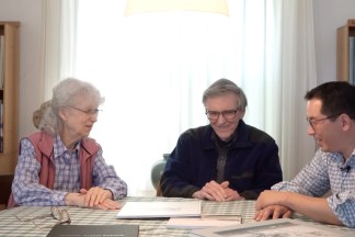 Color screenshot of three figures talking around a table with several books around them