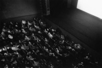 Black and white photograph of theater interior with audience watching screen