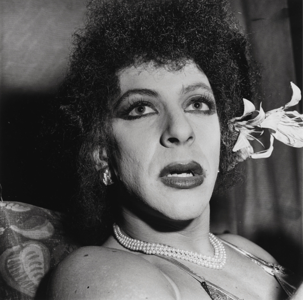 Black and white photograph of a person with curly hair, lipstick, and pearl necklace with a lily flower tucked behind one ear