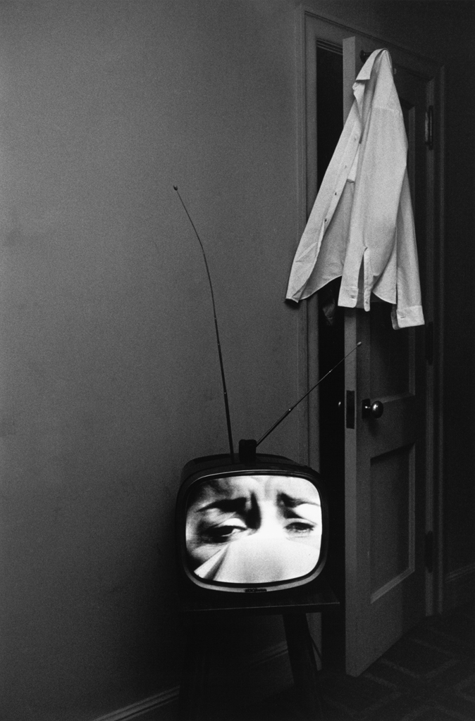 Black-and-white photograph of a television with a crying face on screen and a shirt hanging from an open door
