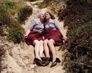 Color photograph of women in matching outfits of lilac tops, red skirts, and black high heels seated cheek-to-cheek on a hillside