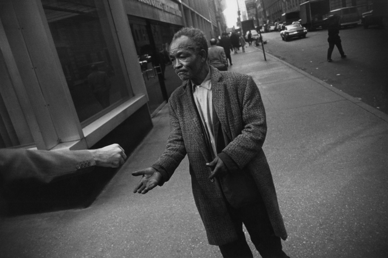 Black-and-white photograph of an outstretched hand making an offering to an older man in a long wool coat
