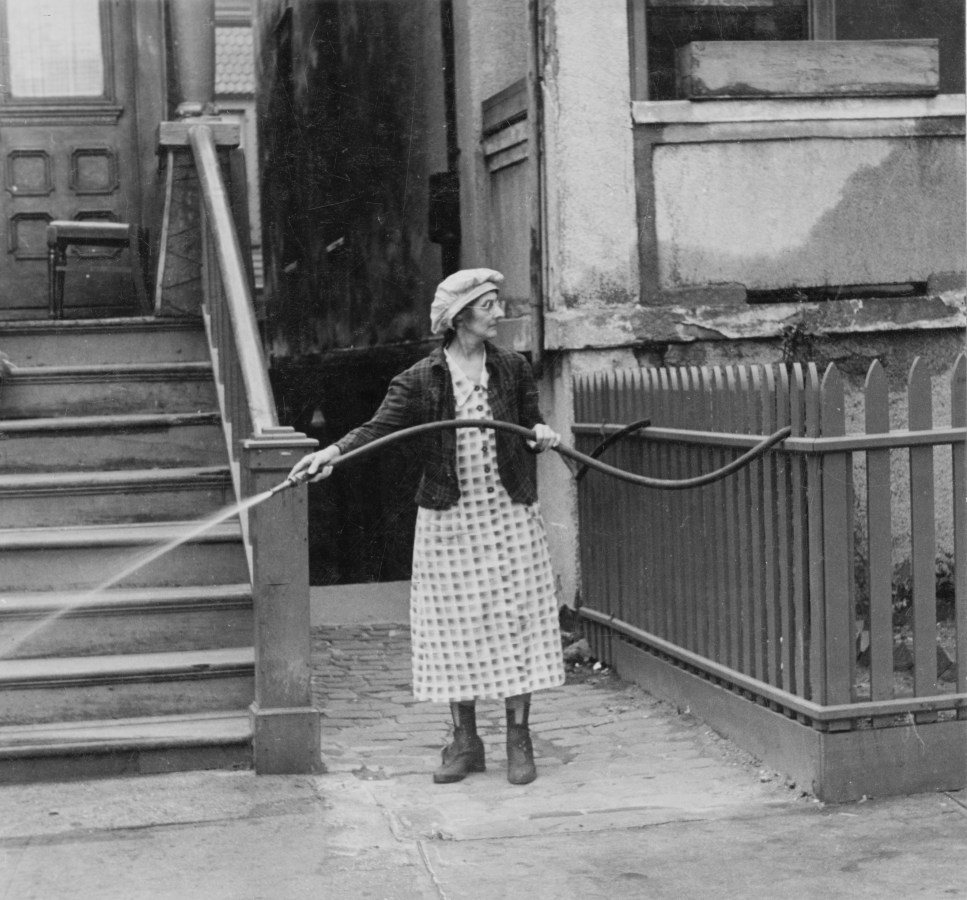 Black-and-white photograph of a woman in front of a brownstone, holding a watering hose.