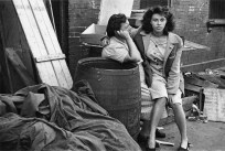 Black-and-white photograph of a young woman looking at the camera seated in the lap of an older woman who is looking away.