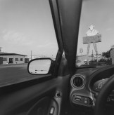 Black and white photograph of a car's dashboard and windscreen in a suburban setting