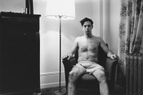 Black and white photograph of a man wearing only boxer shorts seated in an armchair in the corner of a living room