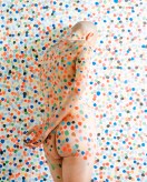 Color photograph of a bald nude figure covered in colorful dots in front of white wall also covered in colorful dots