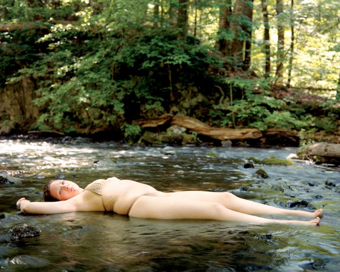Color photograph of a woman in a bikini lying on her back in a shallow rocky creek in a forest