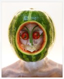 Framed color photograph of a person wearing a hollowed-out watermelon on their head with various vegetables over their face