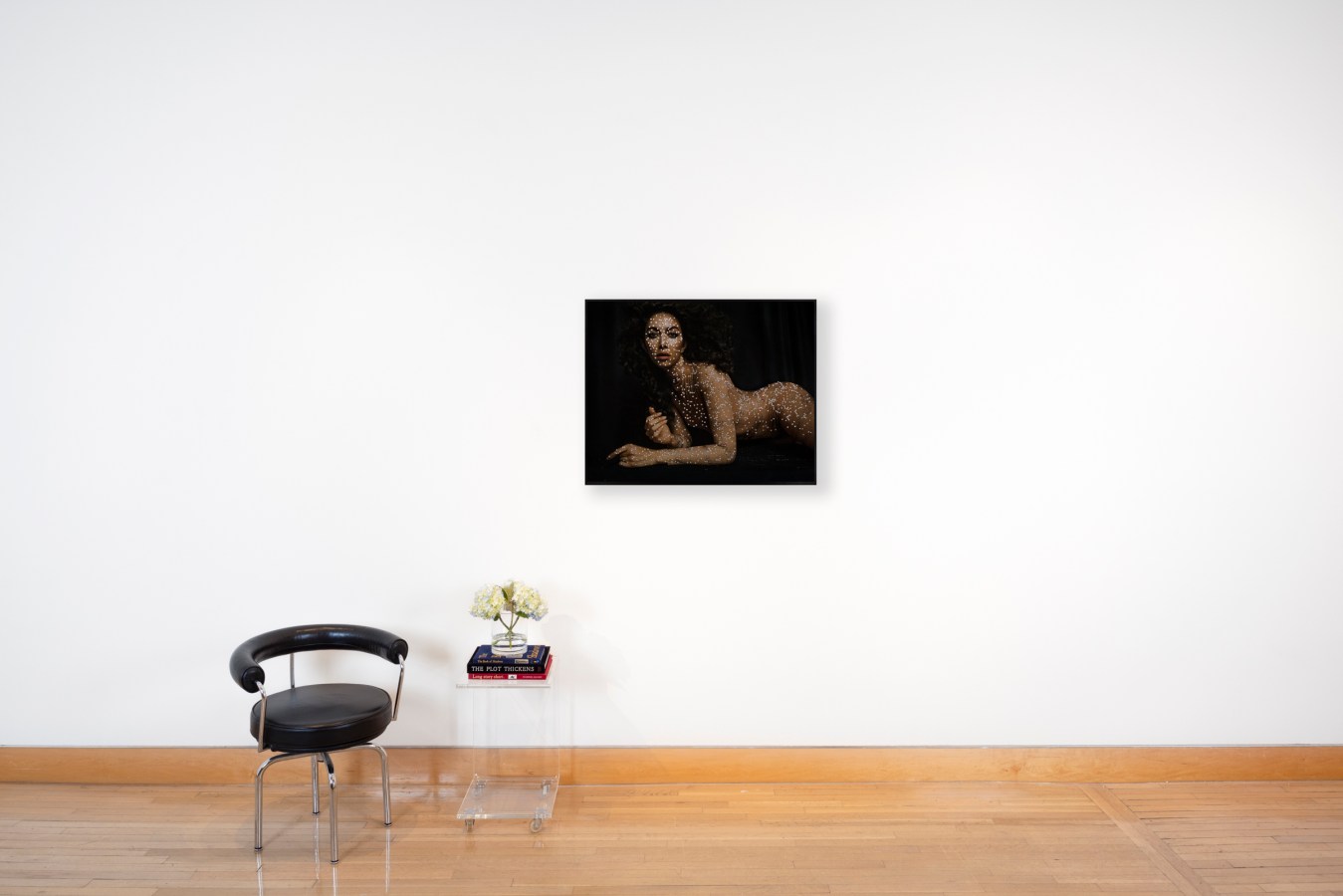 Color photograph of a chair and clear acrylic side table with a color photograph of a nude figure laying and covered in diamonds framed in black on a white wall