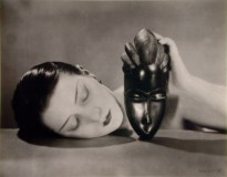 Black and white photograph of a woman laying her head on a table while propping up an African mask in one hand