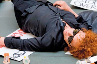 Color image of a figure wearing sunglasses laying on the ground amongst protest signs and empty pill bottles