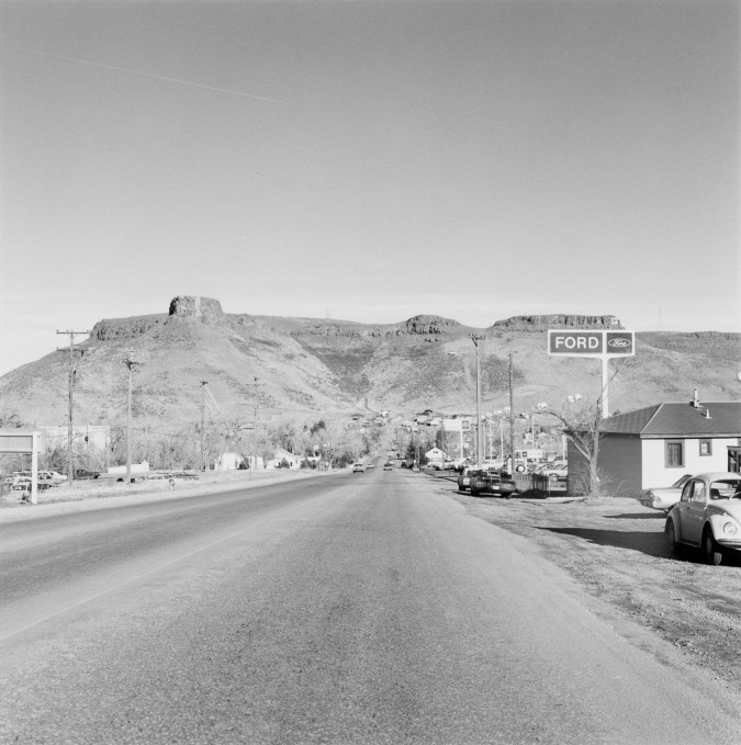 Black and white photograph of a stretch of road with businesses and homes leading to the mountains