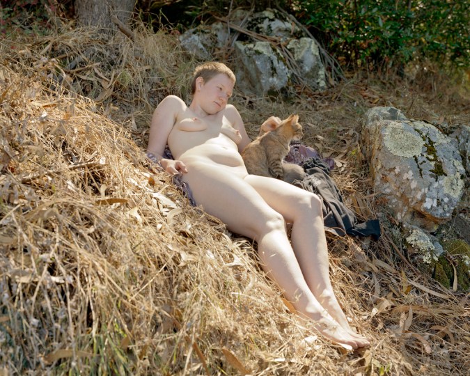 Color photograph of a nude young woman reclining on a grassy hillside petting a cat at her side