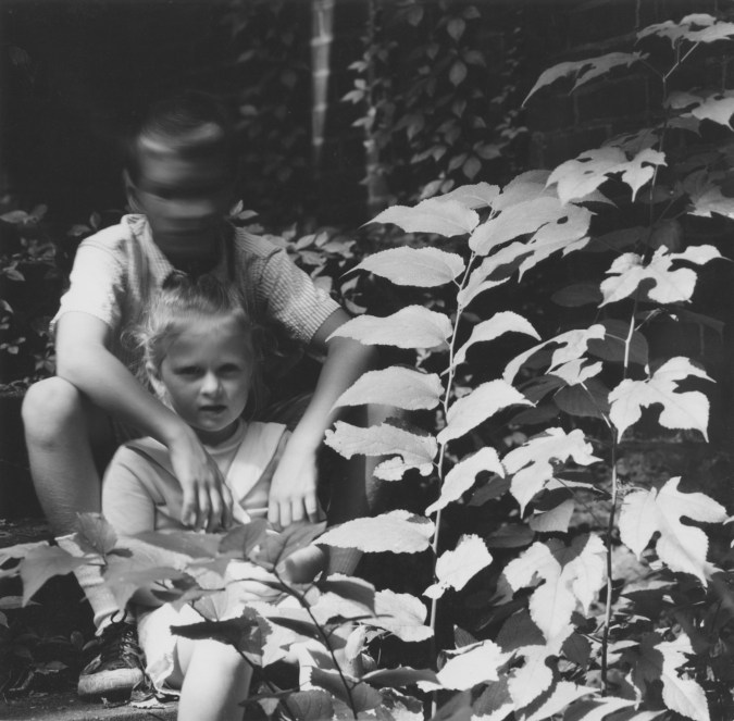 Black and white photograph of a small boy and girl seated near a leafy plant