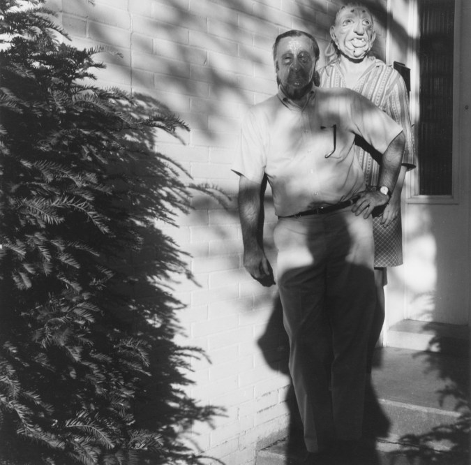 Black-and-white photograph of two people in rubber masks standing on the front steps of a brick home