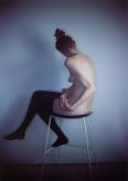 Color photograph of a nude white woman in black stockings, seated ono a stool with her back to the camera.