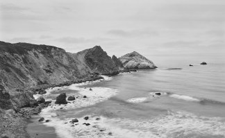 Black-and-white photograph of a cape curving around a bay with waves crashing on a rocky shore