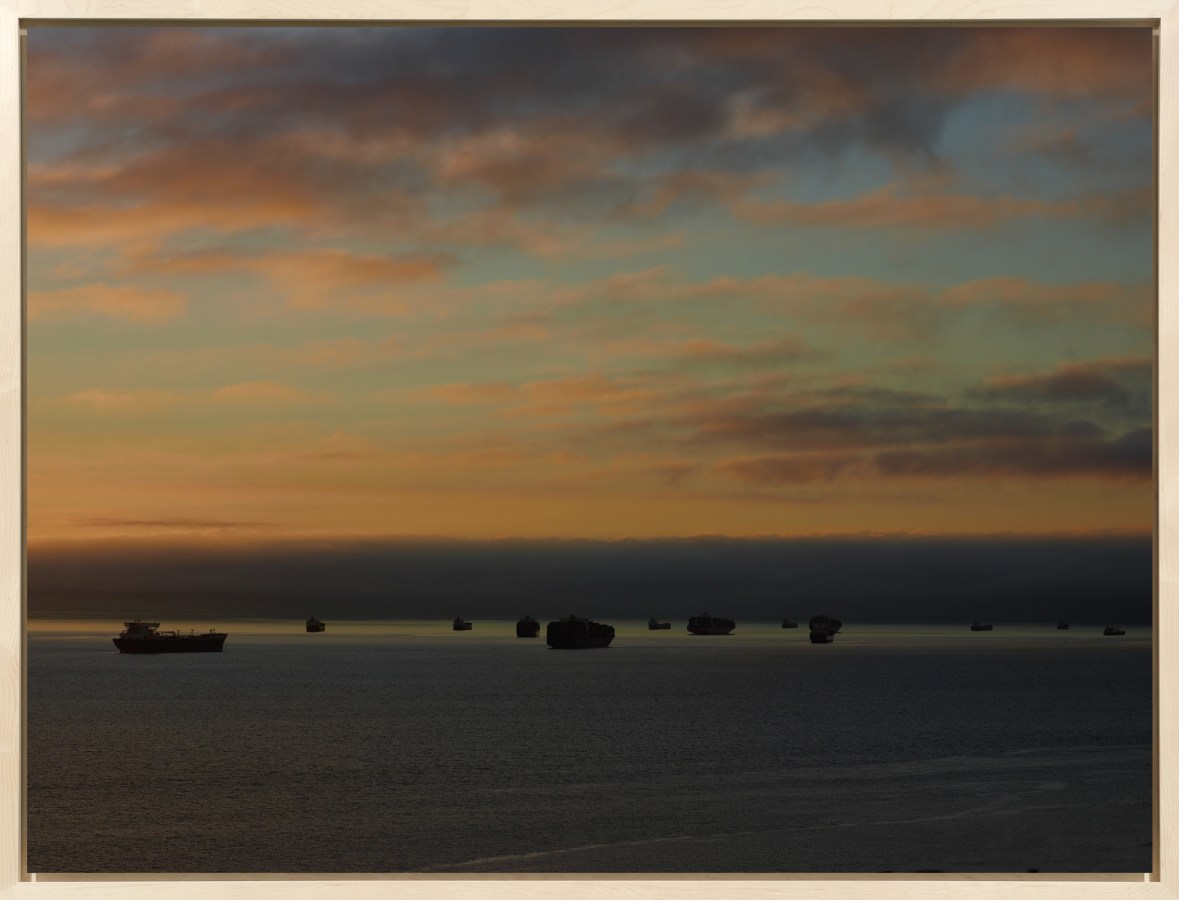 Color photograph of several cargo ships on the horizon during sunrise with pinkish clouds