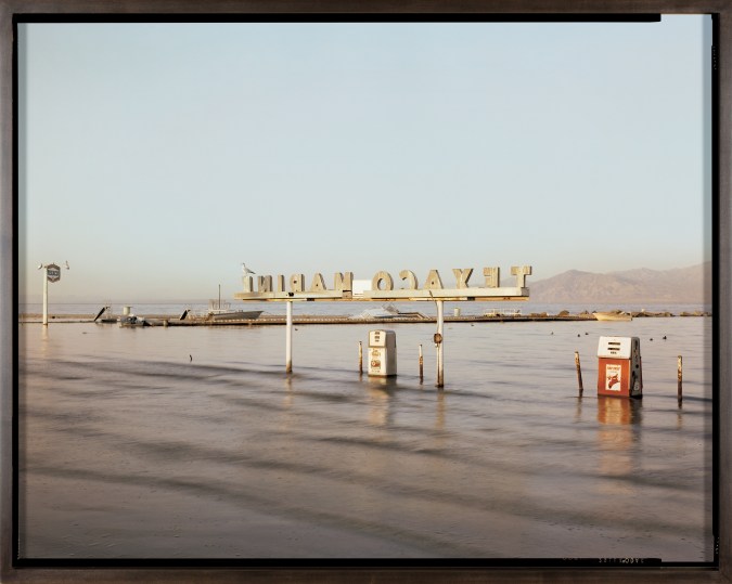Color photograph of a submerged gas station in large body of water with mountain range on the horizon