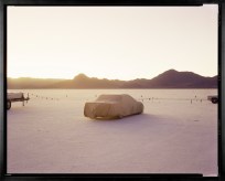 Color photograph framed in black depicting a covered car within the desert with sunlight breaking over mountains on the horizon
