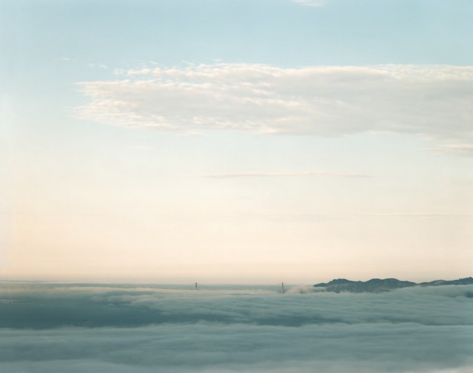 Color photograph of the Golden Gate Bridge on the horizon poking through low fog
