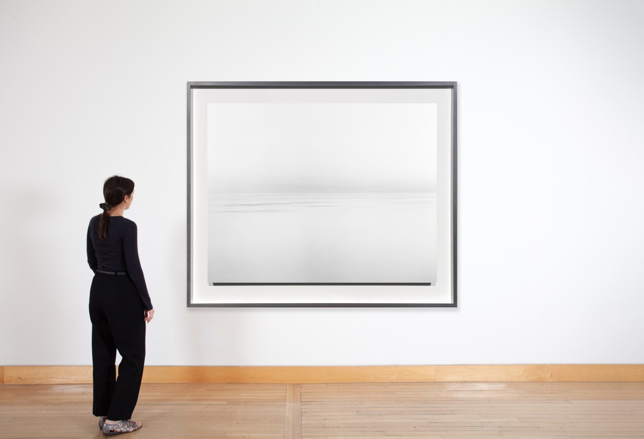 A color photograph of a figure standing in front of a framed black and white photograph of an all white seascape