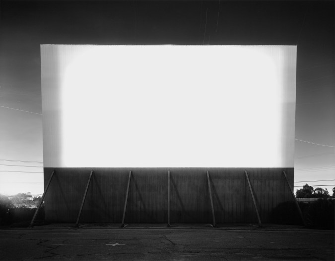 Black and white photograph of an empty drive-in with a blank white screen illuminating the space