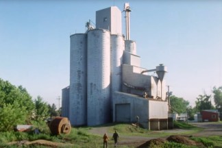 Color video still of a grain silo with two figures walking towards silo