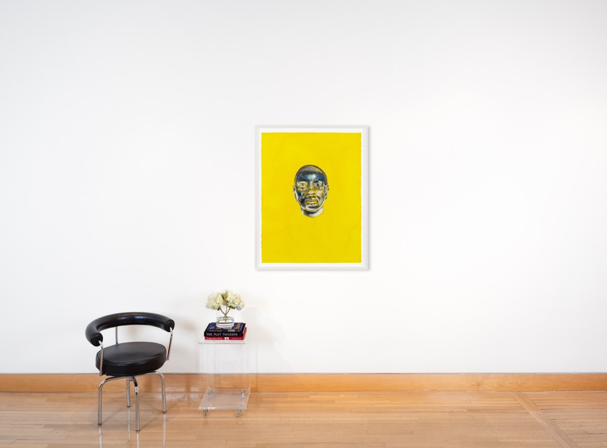 Color image of a chair and clear acrylic table with a mixed media artwork depicting a face in a field of yellow framed in white on a white wall